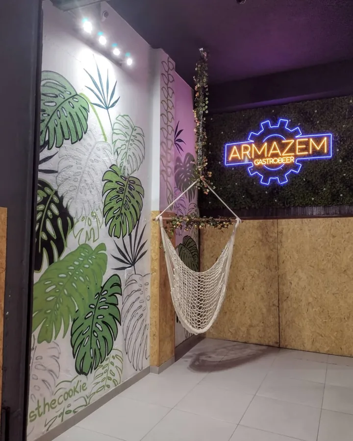 Parede com folhas de costela-de-adão pintadas em tons de verde, ao lado de uma rede e de um letreiro neon