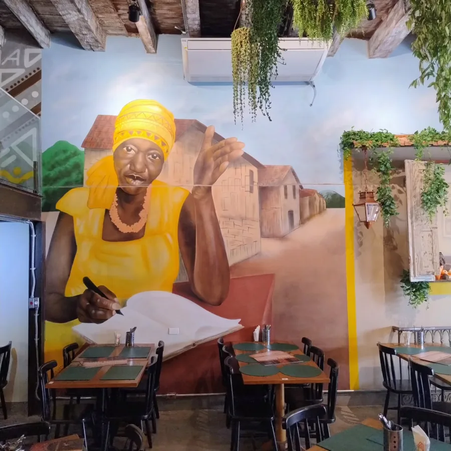 Mural em restaurante: baiana de turbante amarelo escrevendo em um caderno, com casario ao fundo
