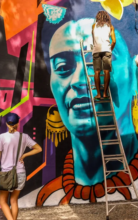 Artista no alto de uma escada pintando um mural colorido de Frida Kahlo em tons de azul, rosa e laranja