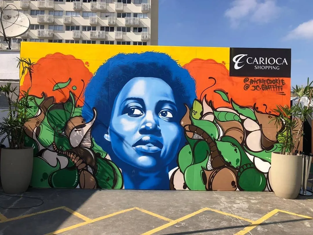 Mural grande em muro externo: rosto de mulher pintado em tons de azul sobre fundo laranja e amarelo, cercado por folhagens verdes