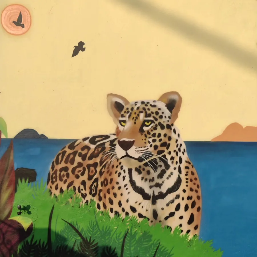 Onça-pintada realista deitada na grama, com lago azul e araras ao fundo, pintada em muro de casa